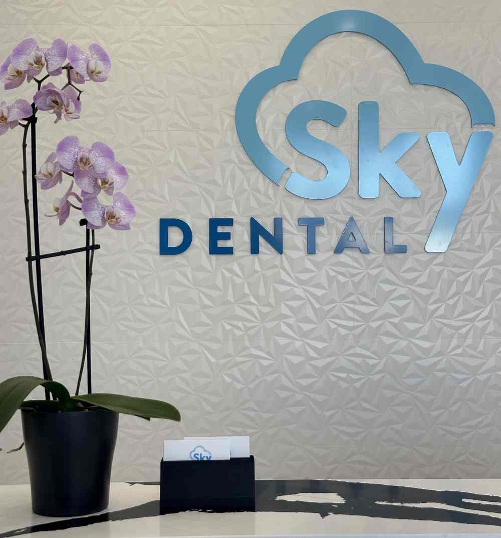  dental clinic desk with the Sky-Dental logo in the background and a purple flower on the front desk. The logo features a stylized sky-blue tooth with a gentle swooping cloud design, symbolizing care and comfort.